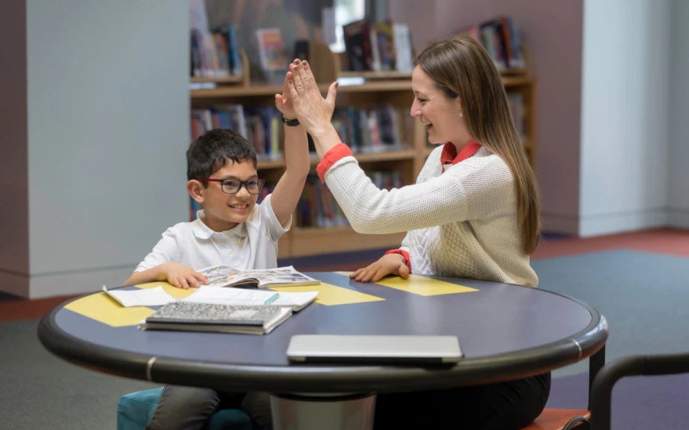 In a classroom with bookshelves in the background, a female instructor in a white jacket high fives her student, who is wearing glasses and a white polo shirt, while they sit at a table with papers and books on top.